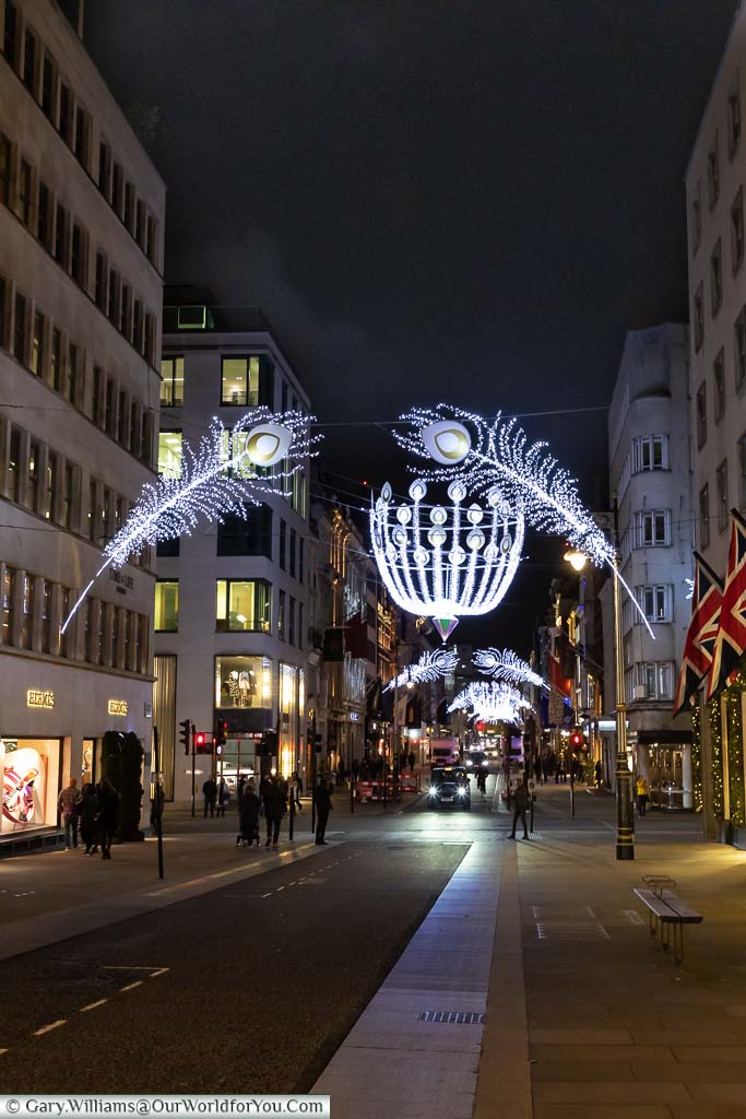 A view down New Bond Street in London’s West End decorated with illuminated silver peacock feathers, and giant baubles above the junctions.
