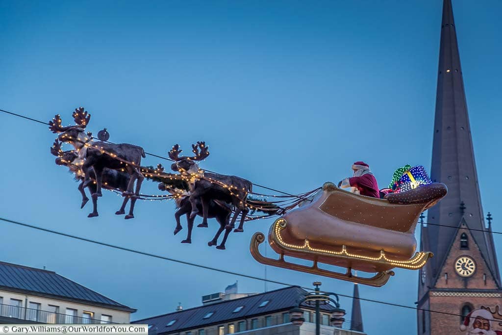 Santa in his sleigh, pulled by his reindeer, soaring above the Hamburg Christmas Market with a church tower in the background.