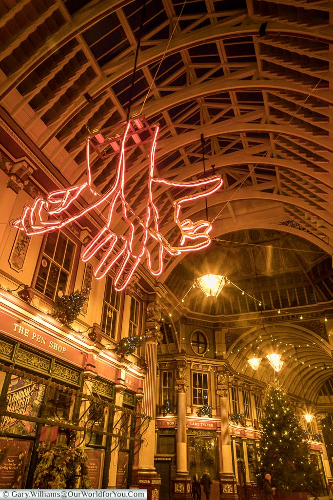 Inside Leadenhall Market at Christmas, looking towards the ceiling of this historic covered market within the City of London. Hanging from the ceiling is an red neon light art installation of 3 hands reaching out, and at the Centre of the market is the Christmas tree.