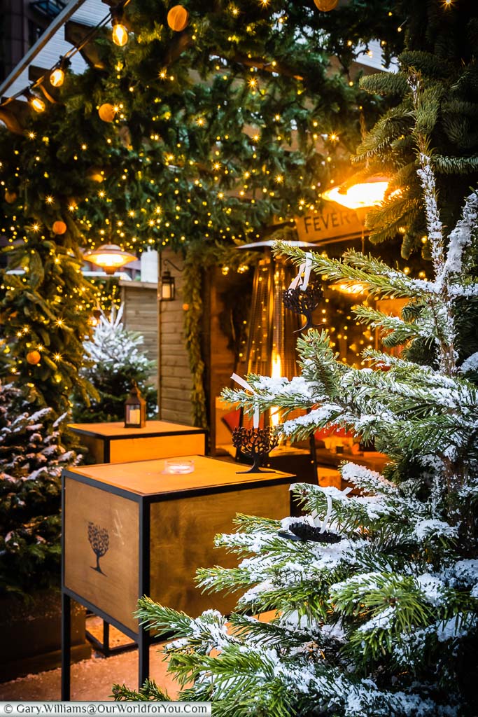 A portrait view of a secluded log cabin the Fever Tree stall at Broadgate's Winter Forest. The scene is edged with a Christmas trees and decorative fairy lights.