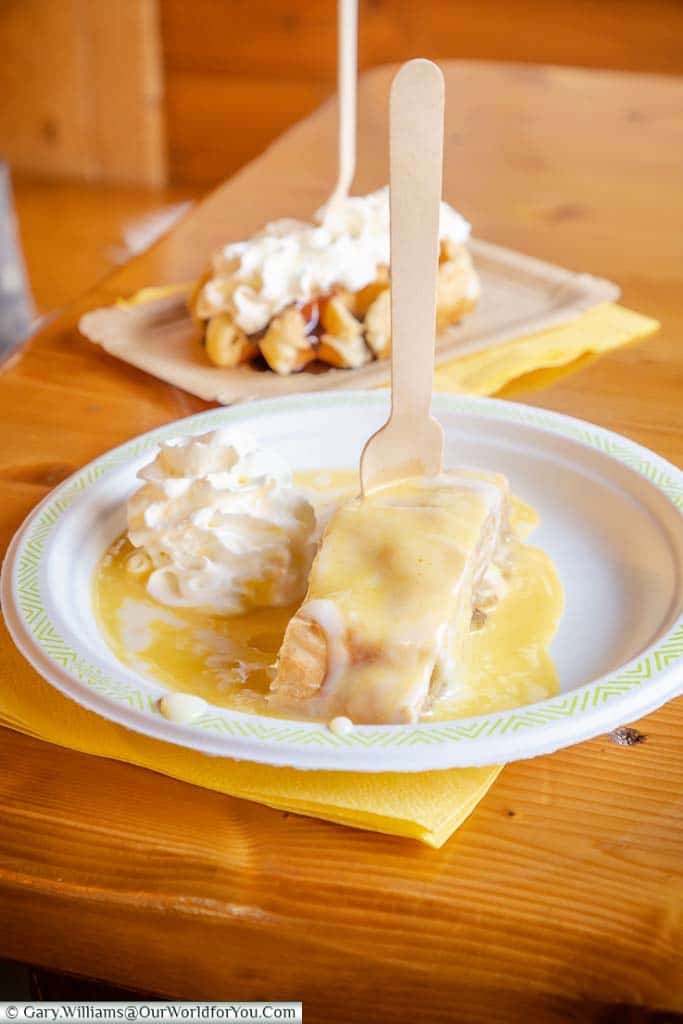 An Apple Strudel & Belgium Waffle on paper plates on a wooden table inside a food hut at Aachen Christmas Markets