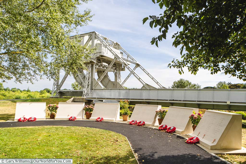 A row of stone monuments, each with a Remembrance wreath, the Pegasus Bridge is in the background.