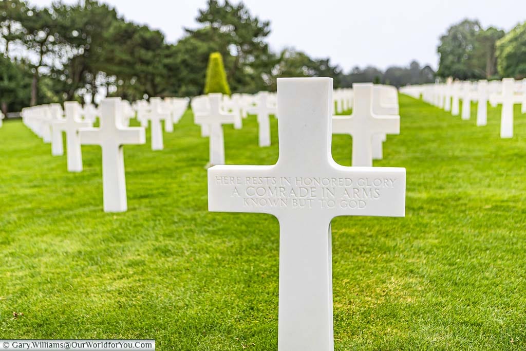A simple white marble cross dedicated to an unknown soldier in front of rows of further headstones in the Normandy American Cemetery