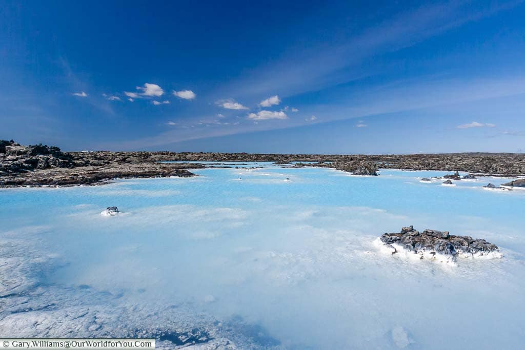 The aqua coloured water of the original Blue Lagoon amongst dark volcanic rocks, caked in mineral deposits where the water comes into contact with them