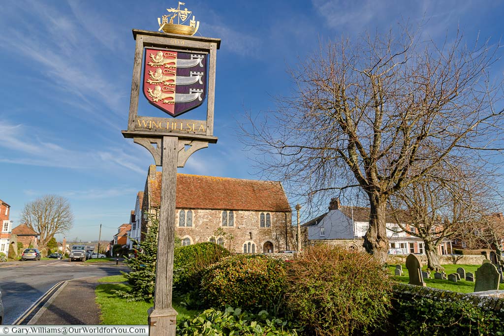 The village sign for Winchelsea, featuring the coat of arms for the Cinque Ports, with the medieval Court Hall in the background.