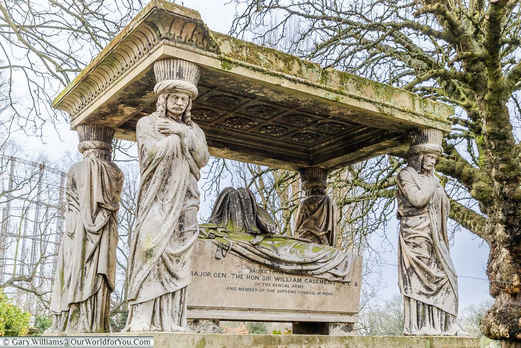 An ornate raised tomb to Major General Sir William Casement, K.C.B. The stone coffin chest is draped with a cloak and bicorn hat. The Canopy is supported by four Indian bearers, each wearing a turban and with his arms crossed.