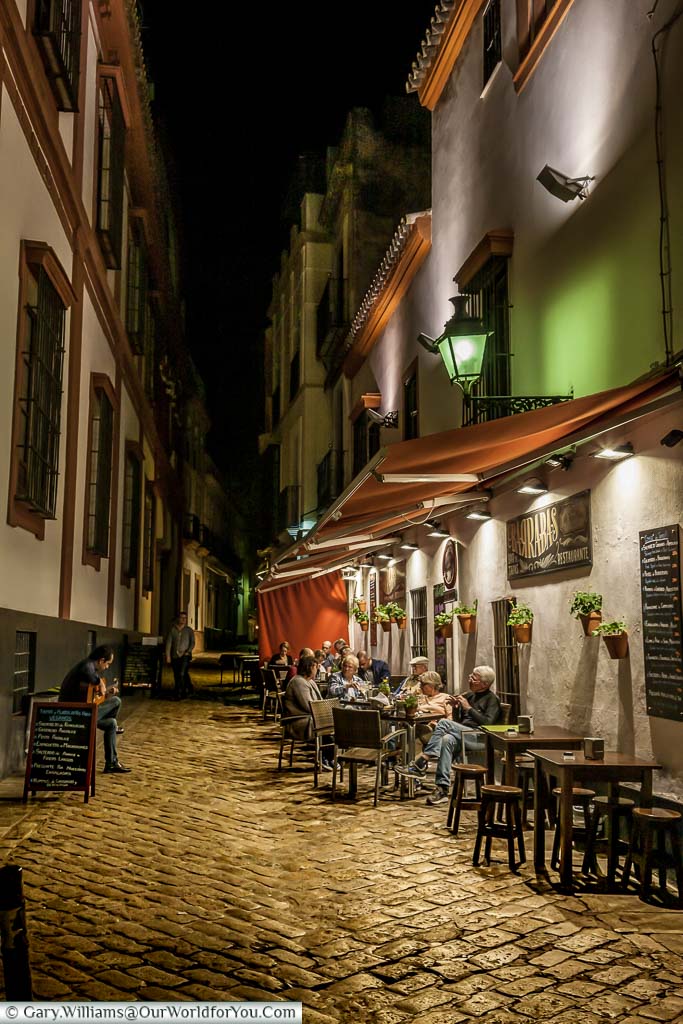 A musician plays a Spanish guitar, in a narrow cobbled lane, in front of a group of people sitting outside a restaurant at night in the Barrio Santa Cruz area of Seville.