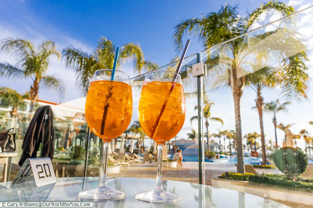 Two glasses of Aperol spritz on a glass table by the palm tree lined pool area of the Olympic Lagoon Resort Paphos