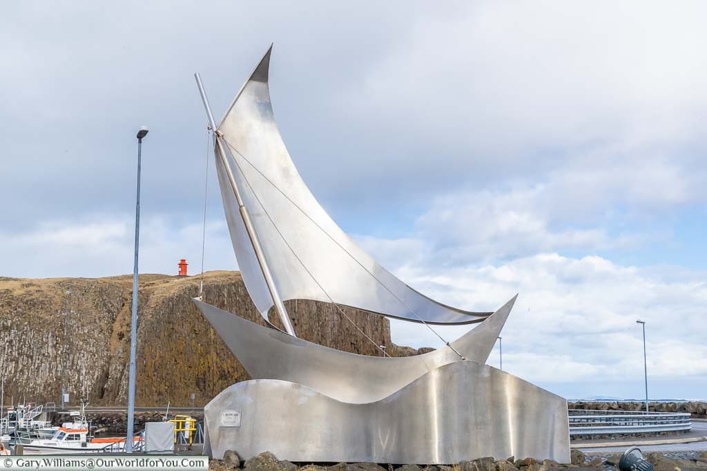 A stainless steel art installation in the town of Stykkishólmur in northwestern Iceland of a sailing boat in front of a red lighthouse atop a rock face in the background.