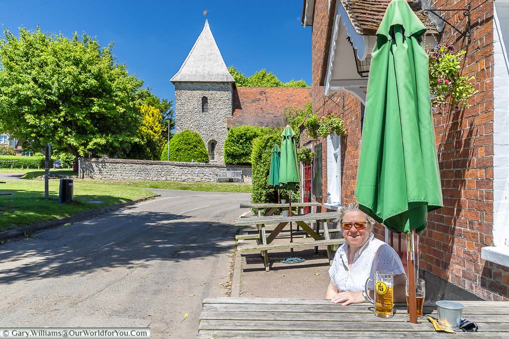 Janis is sitting outside the Swan on the Green in West Peckham with a pint of cold lager on a bright sunny day with St Dunstan's church in the background.