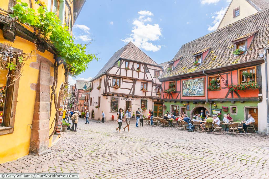 A traditional half-timbered bar & restaurant with tables & chairs spread out into the cobbled square outside in the French town of Riquewihr, on the Alsace Wine Route