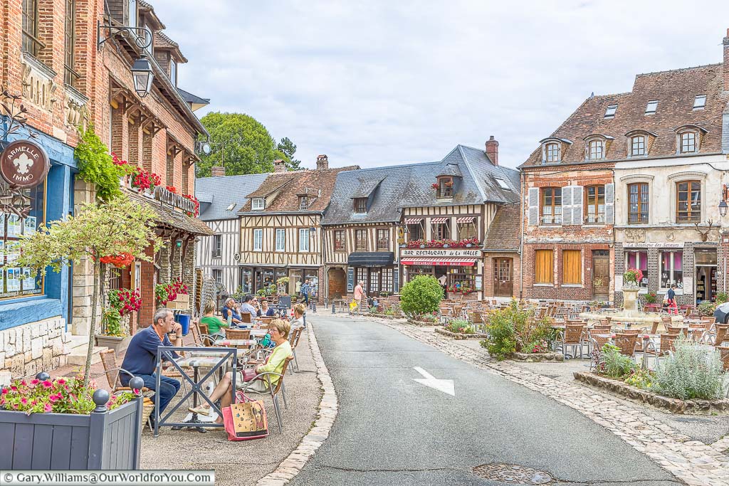 A street scene from the little Normandy village of Lyons-la-Forêt. People are sitting at tables and chairs outside the cafes in this quaint slide of French life.
