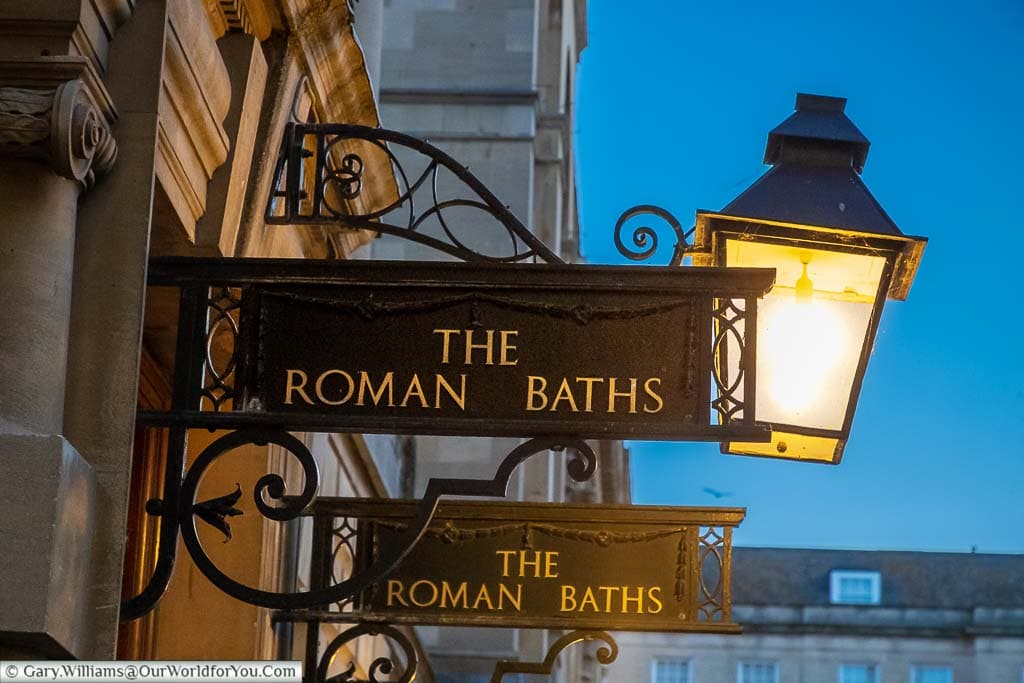 A lantern above two iron signs to the entrance to the Roman Baths in Abbey Churchyard in Bath
