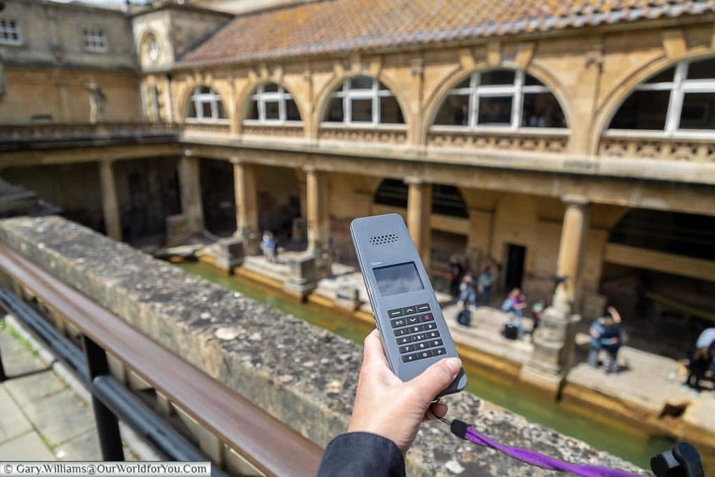 Janis is holding her personal Audioguide in her hand on the upper terrace of the Roman Bath in Bath