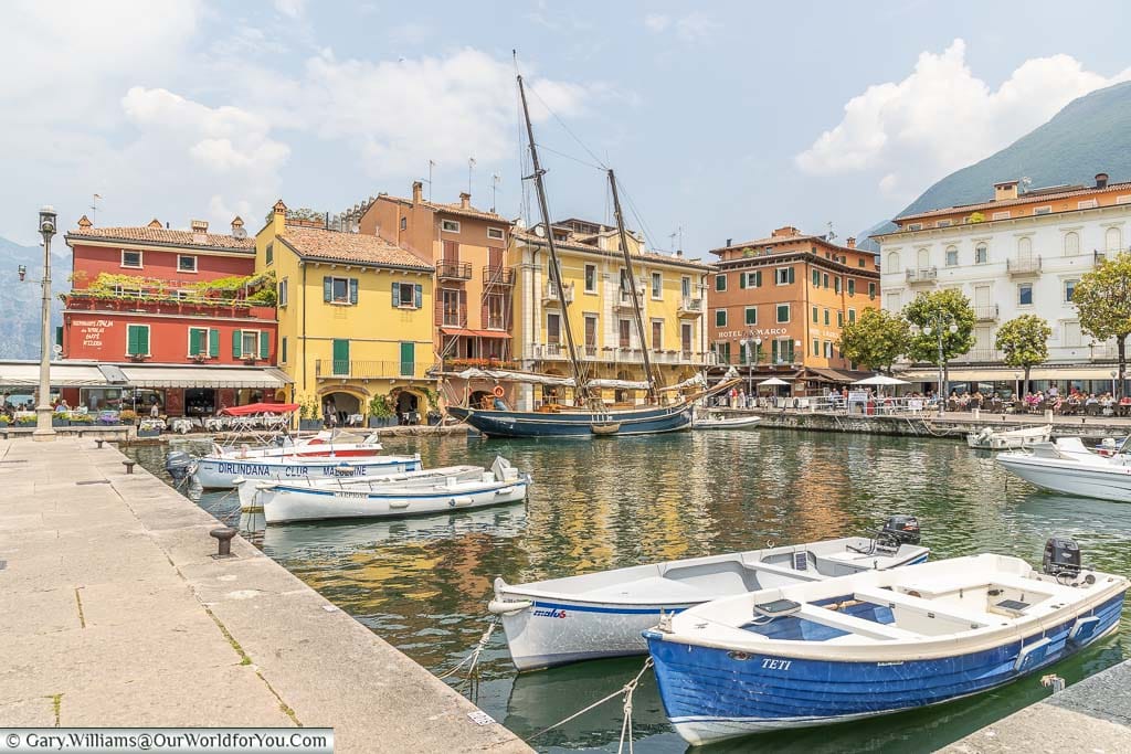 The quaint old harbour of Malcesine lined on two sides with colourful buildings. In the port, there are several small boats and one larger rigged sailboat.