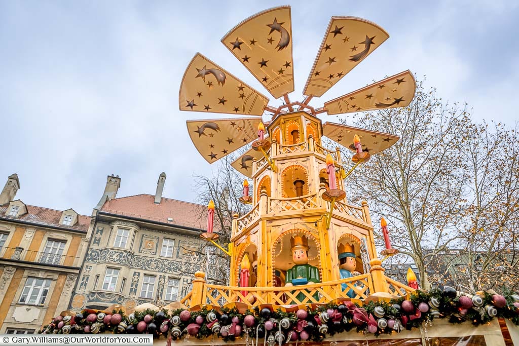A wooden Christmas Pyramid, atop a stall, in the Rindermarkt Christmas markets, against the backdrop ornately decorated buildings that line the square.