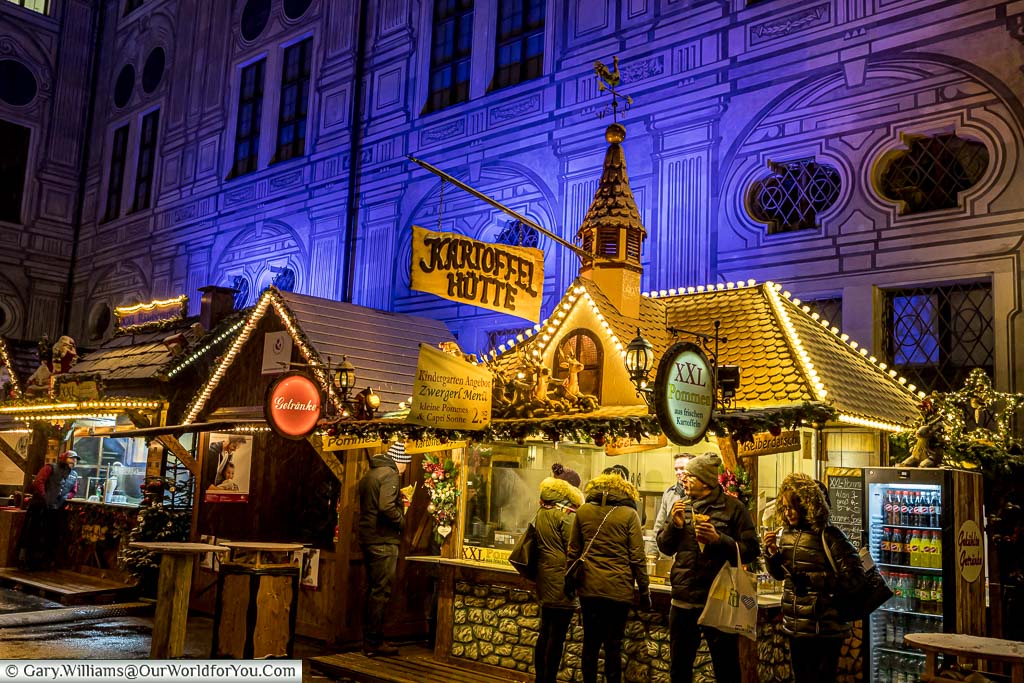 A collection of food stalls in the Munich Residenz Christmas Market with the one in the foreground labelled as the Kartoffel Hütte. The backdrop is blue lit buildings with faux facades.