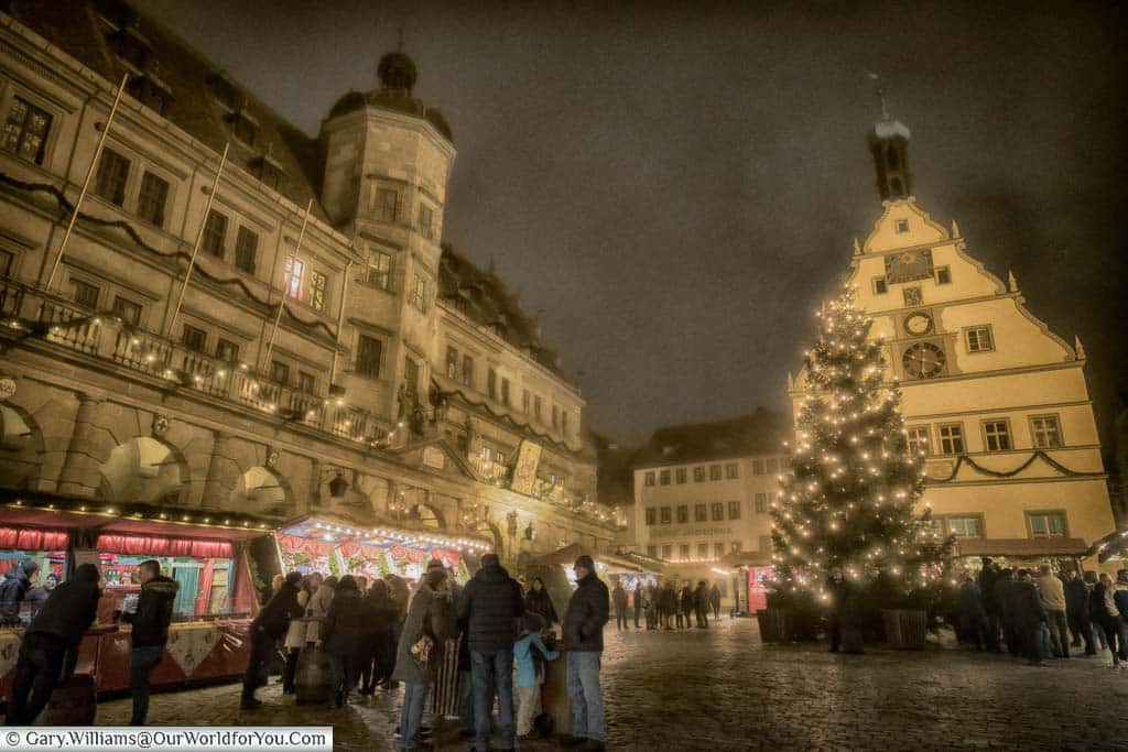 The Marktplatz of Rothenburg ob der Tauber on a misty evening with groups of people huddled together. There are a few stalls on the left in front of the Rathaus, and the Christmas tree to the right in front of another historic building.