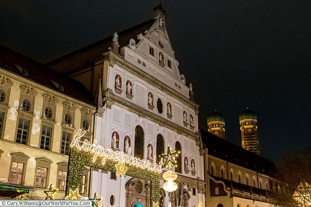 A lit sign welcomes you to the Kripperlmarkt Christmas market, against the backdrop of the historic buildings that line the Neuhauser Straße with the towers of Munich Cathedral visible in the background.