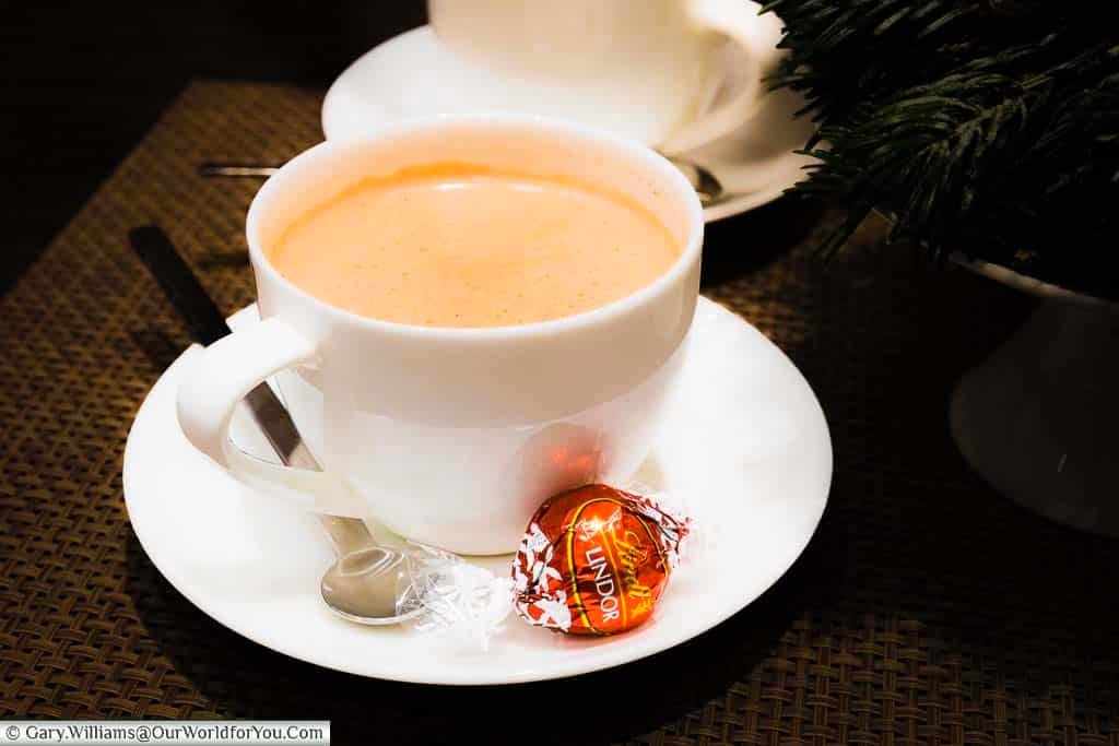 A white china cup full of foaming hot chocolate with a Lindt chocolate on the side.