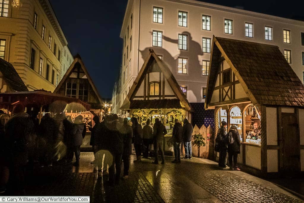 People huddle together stalls in the Medieval Christmas market on a drizzling evening with the silhouette of the statue of Maximilian I reflected on the building in the background.