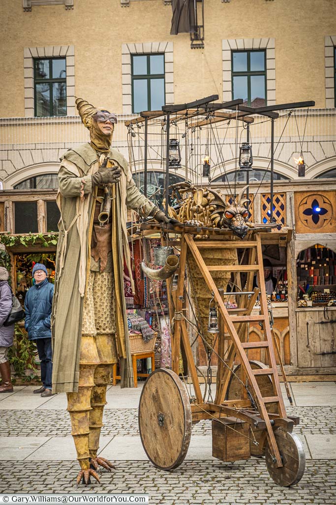 A costumed performer on stilts alongside a cart holding a wooden, fire breathing, dragon puppet in the Medieval Christmas market.