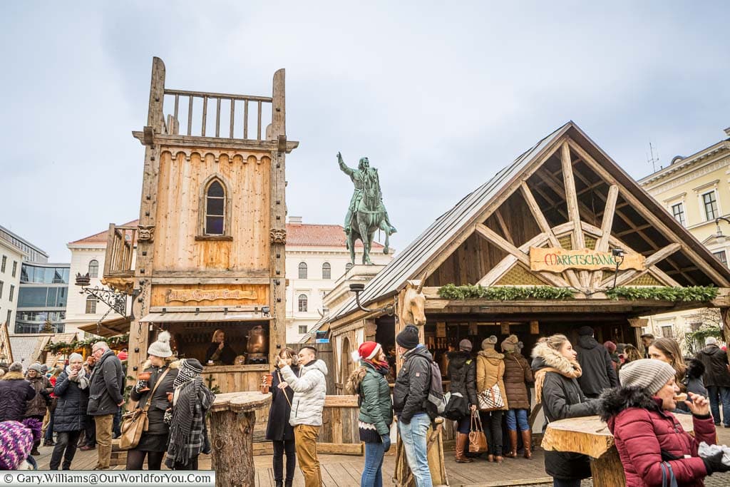 Groups of people in front of the wooden market stalls of the Medieval Christmas market around the statue of Maximilian I in Wittelsbacherplatz.