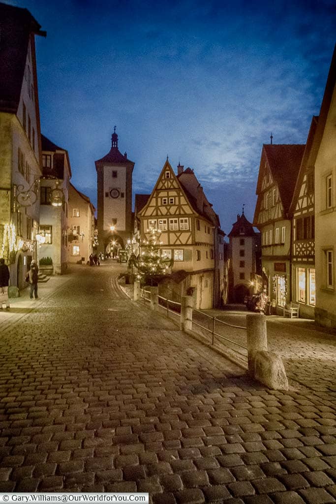 A view of a split in the cobbled lane leading from Rothenburg og der Tauber main town square to one of the gate towers at dusk under a blue sky.