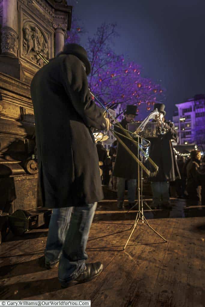 A three-man brass band, wearing long wool coats and top hats, playing around the statue in the centre of Alter Markt in the centre of cologne's christmas markets at night.