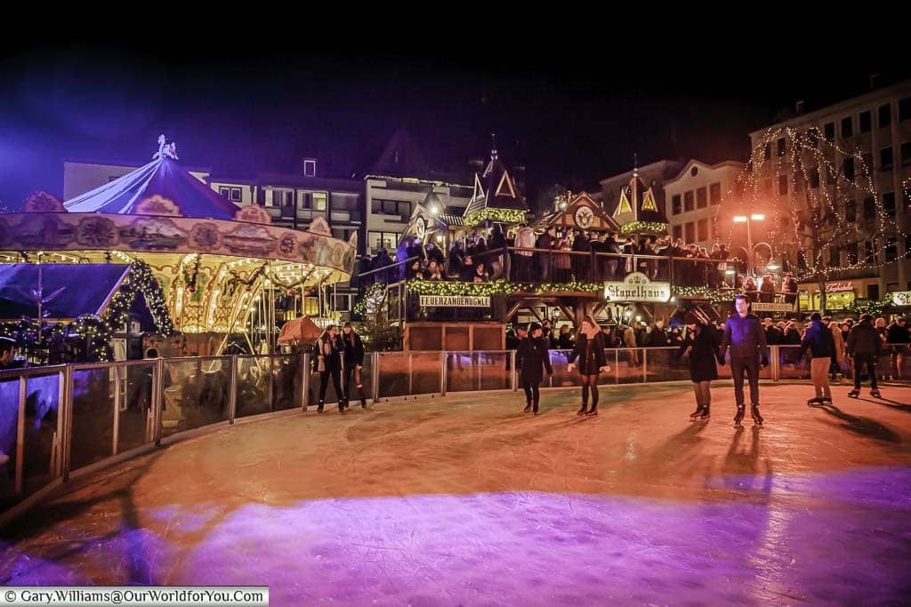 One of the circular ends to the ice rink in Cologne, lit at night, overlooked by a carousel and the two-storey drinks cabin.