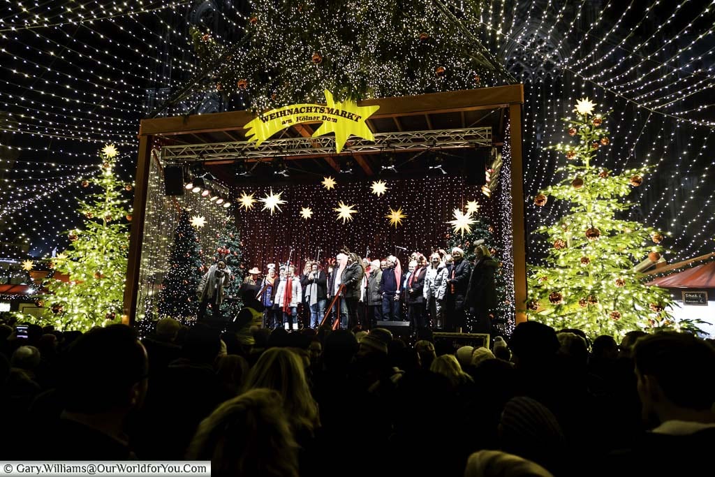 Crowds in front of a carol concert on stage in the cologne's Dom Christmas Market, under a blanket of fairy-lights