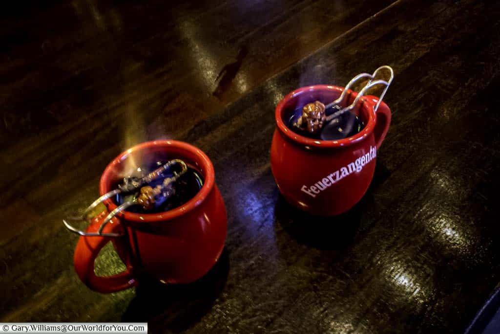 Two red mugs of flaming feuerzangenbowle with rum-soaked sugarloaf dissolving into the liquid below on the counter of a drinks stall on cologne's christmas markets.