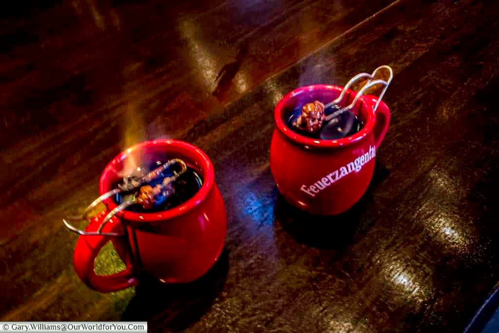 Two red mugs of flaming feuerzangenbowle with rum-soaked sugarloaf dissolving into the liquid below on the counter of a drinks stall on cologne's christmas markets.