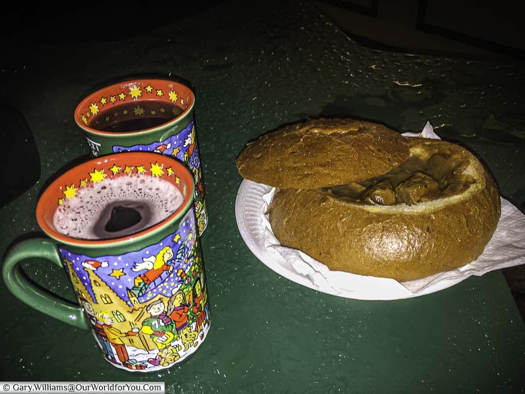 A large scooped out roll filled with Gulaschsuppe next to two mugs of steaming glühwein from the Cologne Christmas Markets