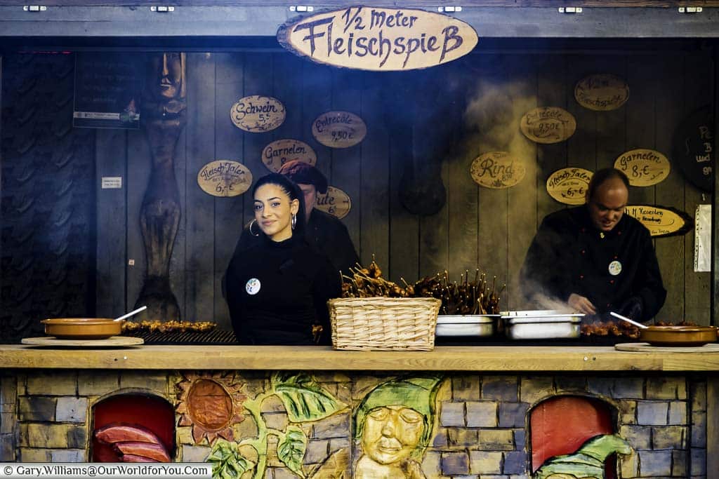 A smiling young lady awaits a customer at the kebab stall, while in the background, you can see the cooks preparing the skewered kebabs over hot coals.