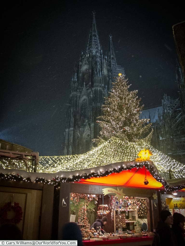 A stall in front of Cologne's main Christmas tree, in the centre of cologne's christmas market, next to the twin spires of the Dom as it snows.