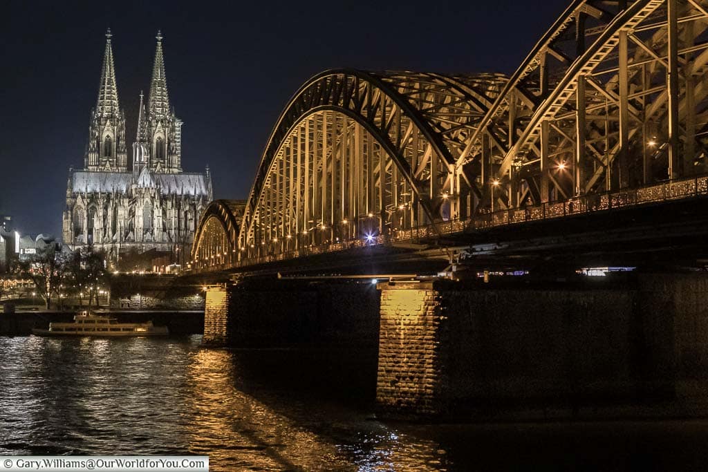 The iconic evening view of the Dom and Hohenzollernbrücke. The cathedral is lit by white lights and the arched iron railway bridge is lit by orange lights With the River Rhine flowing underneath.