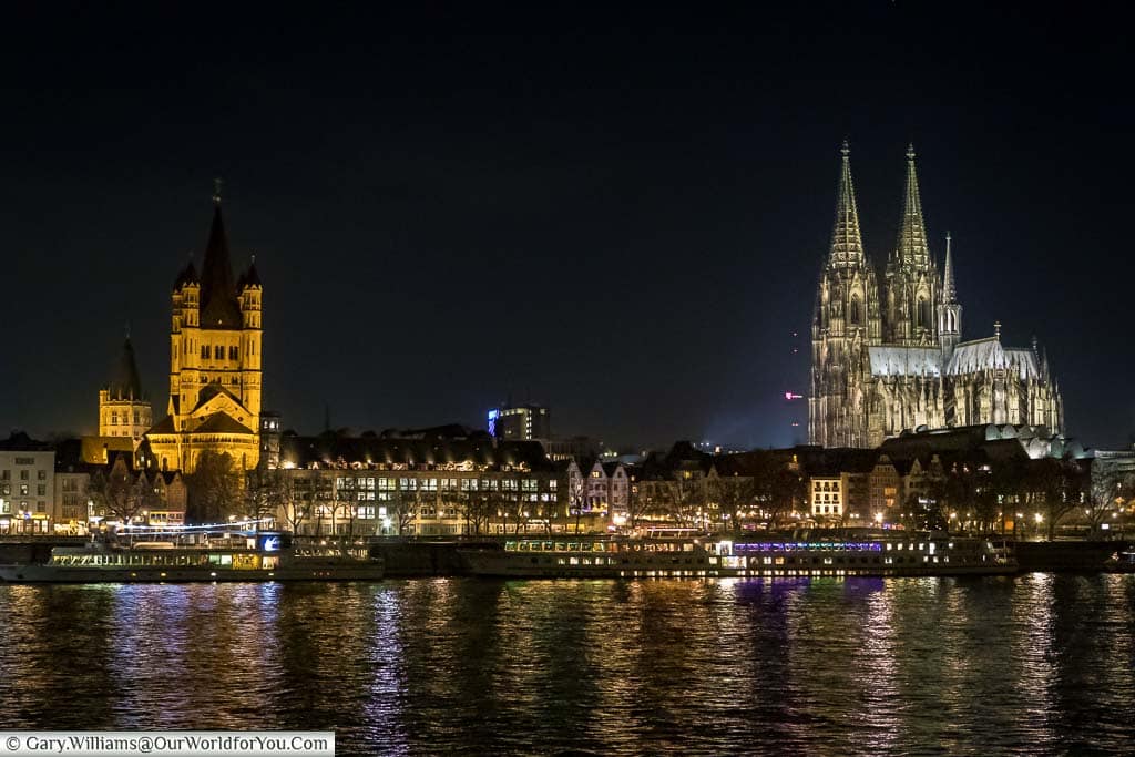 A view across the River Rhine at night with Cologne cathedral dominating the skyline on the right in Saint Martin's church dominating the skyline on the left. Pleasure craft line the edge of the riverbanks are waiting to take passengers on on an evening tour