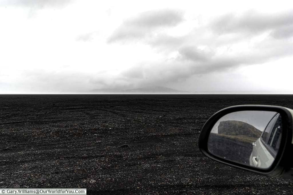 An almost black and white image of the black beach & cloudy skyline from the window of our 4x4 with the only colour from a rock formation in the side mirror