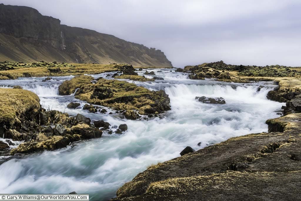 River rapids flowing through the Icelandic countryside next to an escarpment.