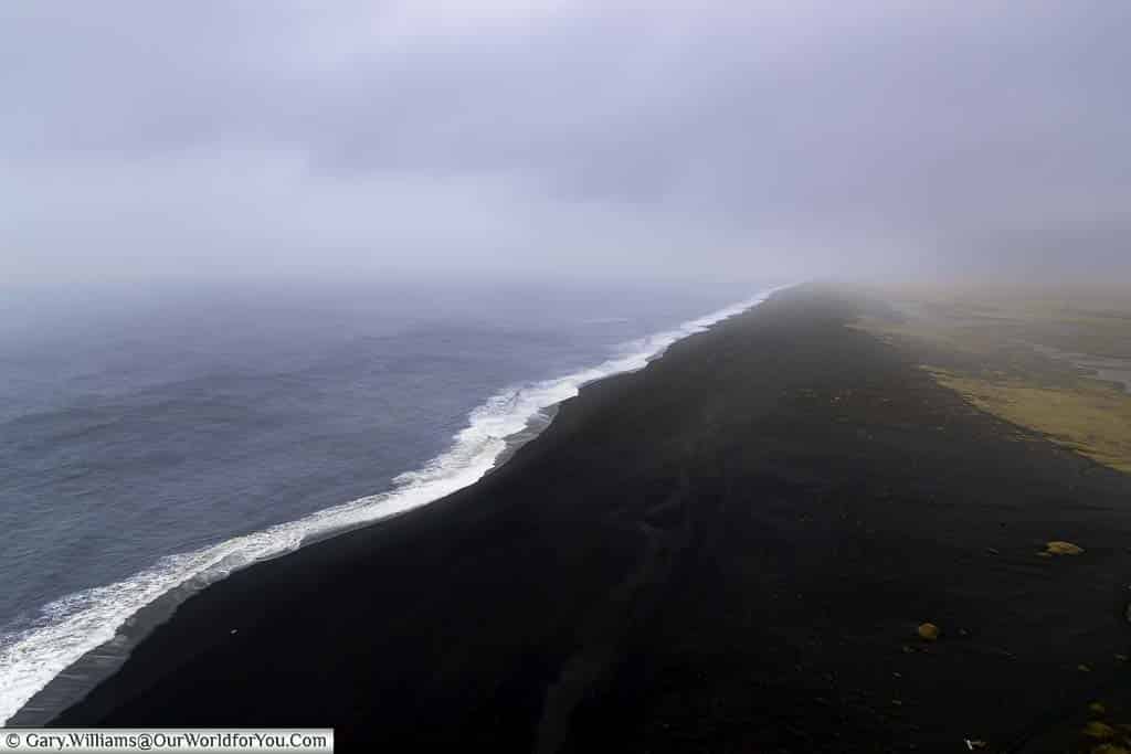 A view from on high of the black beach stretches into the distance where the mist meets the land. The beach is separated from the dark waters of the Atlantic Ocean by a line of white breaking water.