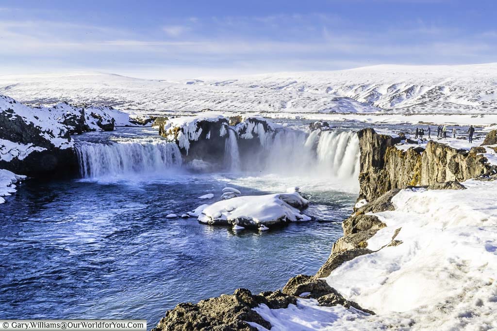 The horseshoe-shaped Goðafoss waterfall against a backdrop of snowy white mountains.