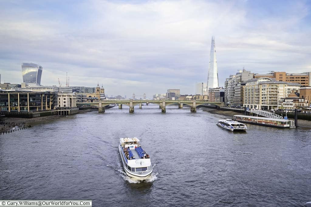 A tourist boat passing under the Millennium Bridge as we look east to Southwark Bridge with The Shard skyscraper in distance.