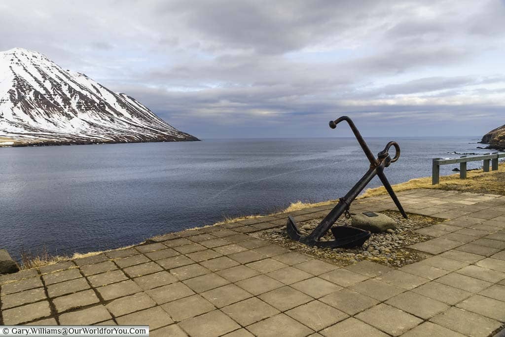 An iron anchor memorial on the pavement next to the water's edge just outside Ólafsfjörður, Iceland