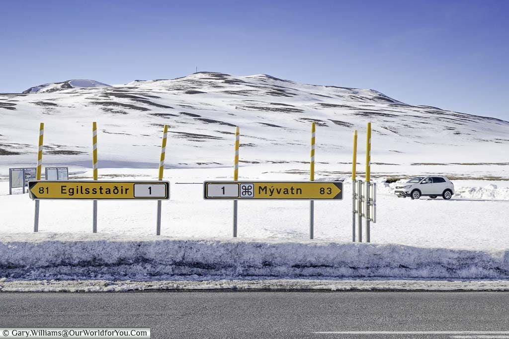 Two yellow road signs on iceland's route one, one pointing to Egilsstaðir 81km away, and the other to Mývatn 83km in the opposite direction, against the backdrop of white mountains of eastern iceland