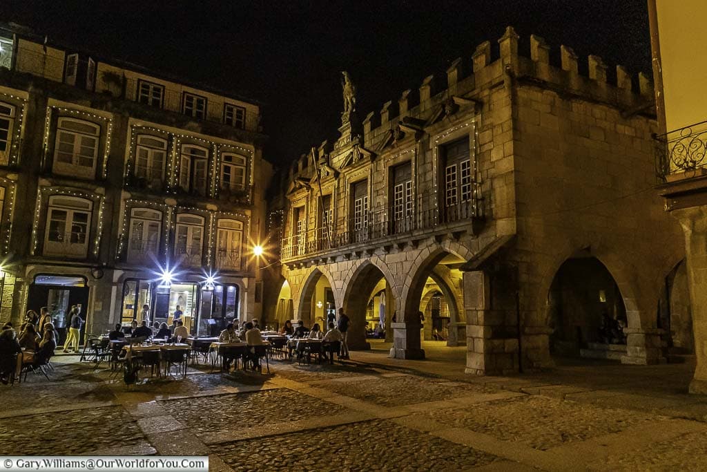 A lit square after dark in Guimarães, Portugal where diners sit at restaurants lining the square and others walkthrough the historical old town.