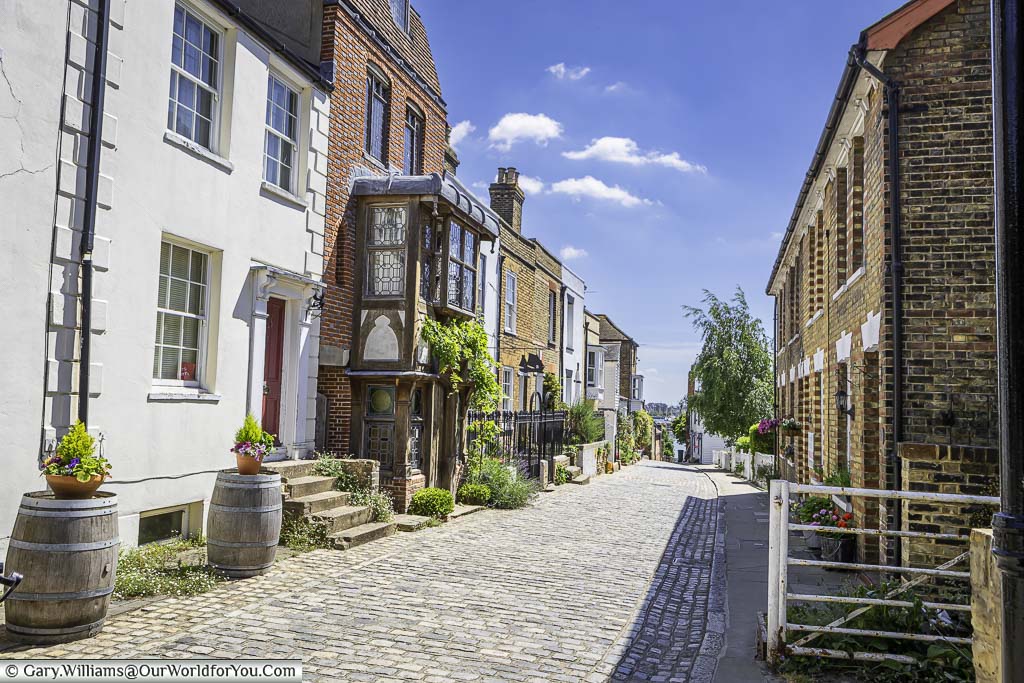 The cobbled high street in Upnor, lined with historic brick-built houses, leading towards the River Medway on a sunny day.