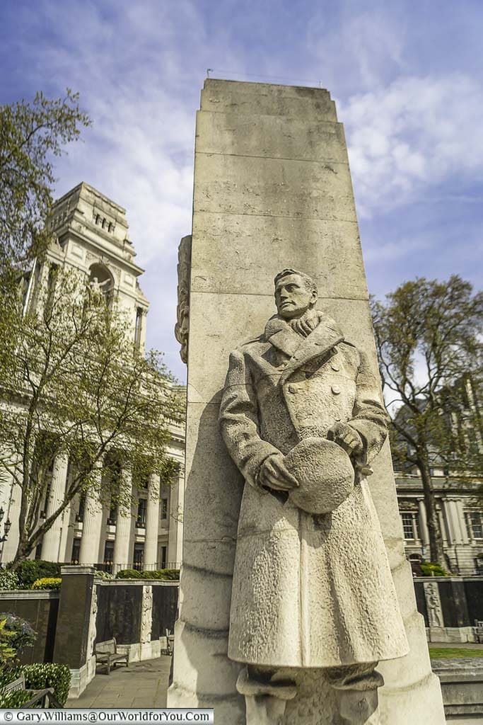 A sandstone statue of a naval officer, wrapped up warmly against harsh conditions, looking into the distance. In the background is Trinity house, a neo-classical designed piece of architecture on the edge of Tower Hill Memorial Gardens