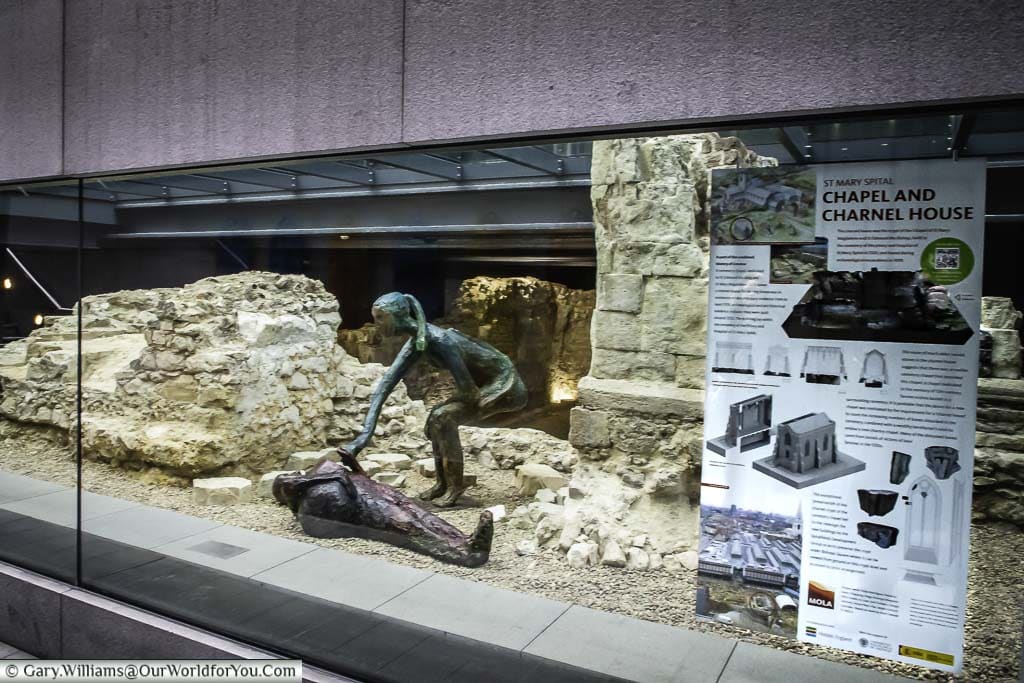 Looking through a large glass panel to the ruins of a 13th-century dwelling in Spitalfields, know as Charnel House, with a couple of bronze sculptures represent the residents to provide scale.