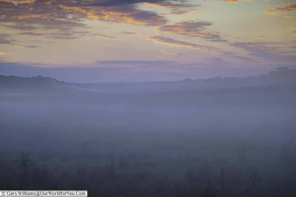 Mist hangs over the valley that used to once be a river bed, with a golden sky, tinged with purples before the sun rises.